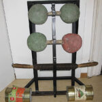 Muckibude mit schwarzes Eisengestell, auf dem drei selbstgebaute Hanteln abgelegt sind, mit davorliegender Hantel aus einem schwarzen Stab und zwei mittelgroßen Dosen, als Objekt im Strafvollzugsmuseum ausgestellt