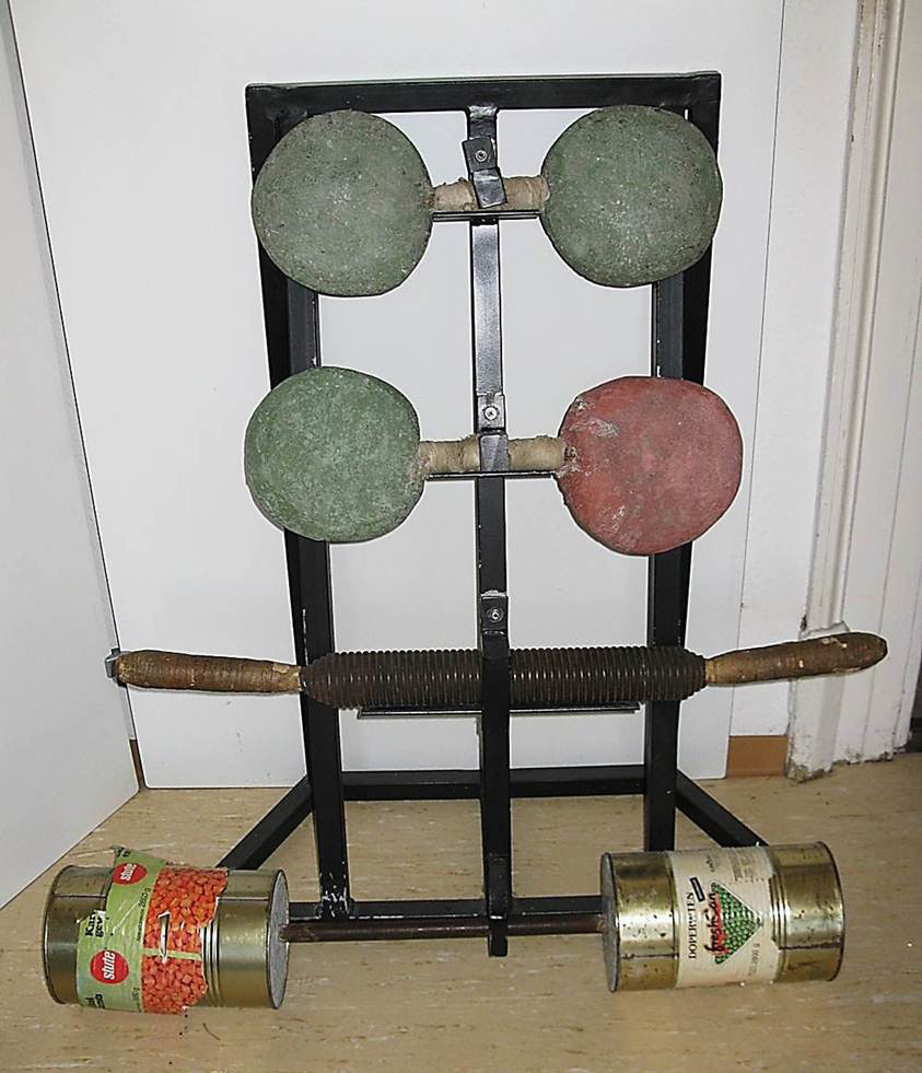 Muckibude mit schwarzes Eisengestell, auf dem drei selbstgebaute Hanteln abgelegt sind, mit davorliegender Hantel aus einem schwarzen Stab und zwei mittelgroßen Dosen, als Objekt im Strafvollzugsmuseum ausgestellt