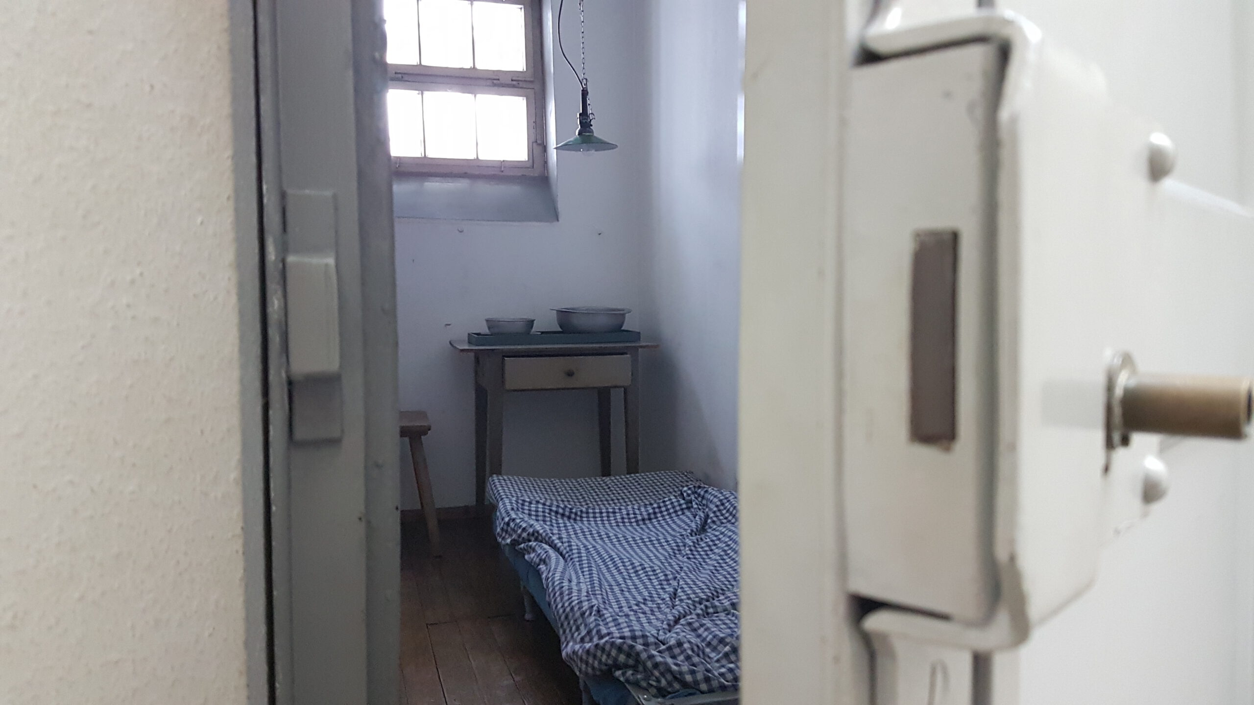 Blick durch eine halb geöffnete Zellentür in eine Haftzelle mit hochklappbarem Bett mit blau-karierter Bettwäsche, einem Holztisch mit zwei Schüsseln und einem Holzstuhl