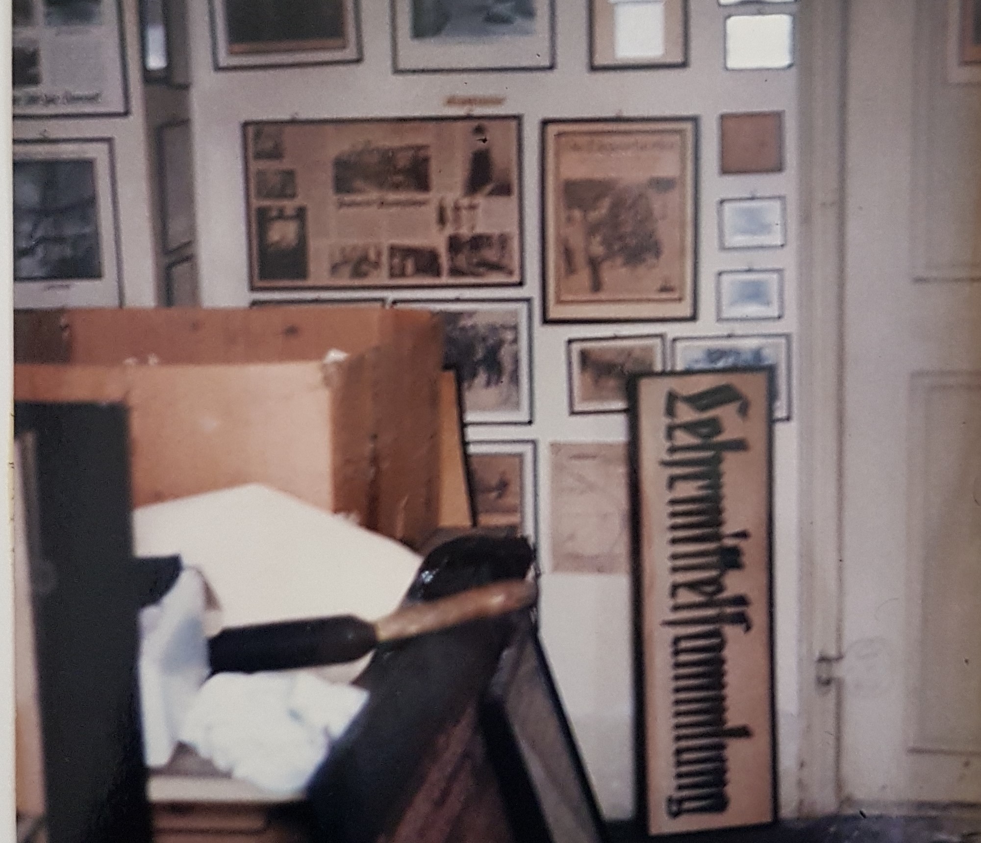 Verschwommener Bildausschnitt auf dem ein Raum des Strafvollzugsmuseums mit links einem mit Kisten und Gegenständen vollgestellten Tisch zu sehen ist, der in einiger Entfernung zu einer mit Bildern vollgehängten weißen Wand steht, an die ein rechteckiges Schild mit der Aufschrift "Lehrmittelsammlung" lehnt