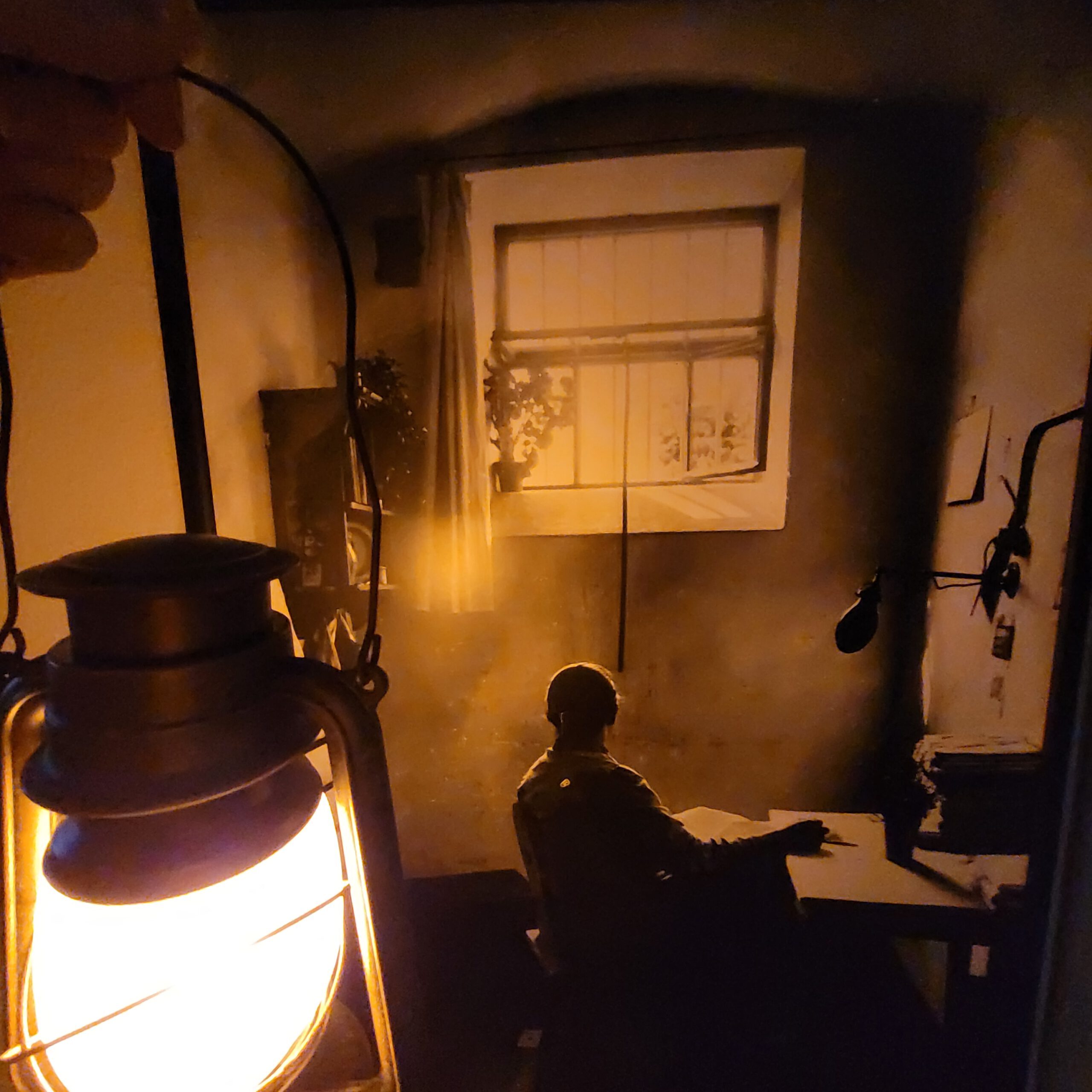 eine Person beleuchtet mit einer Laterne ein Bild, dass einen Gefangenen in einer kleinen Zelle aus den 1930ern zeigt, wie er an seinem Tisch sitzend sehnsüchtig hinauf zum Fenster blickt