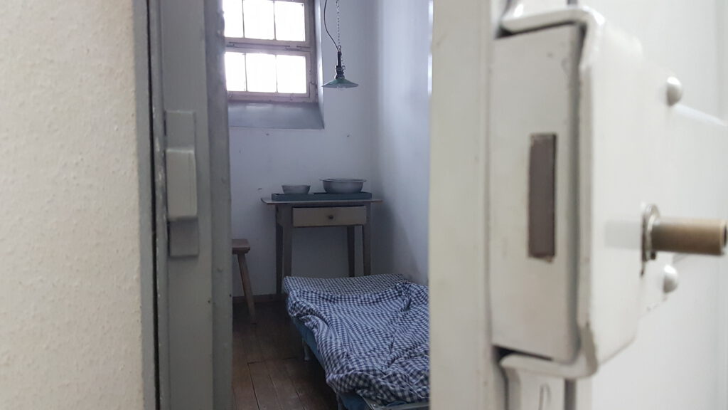 Blick durch eine nur einen Spalt geöffnete weiße lackierte Stahlzellentür in die Einzelhaftzelle im Zustand von 1928, mit Hochschließbaren Bett, darauf die typisch weiß-blau karrierte Bettwäsche, dahinter ein kleiner Holztischmit Schublade, darauf zwei Blechschüsseln, daneben ein Schemel, eine einfache Hängelampe, Licht fällt durch das vergitterte Fenster.