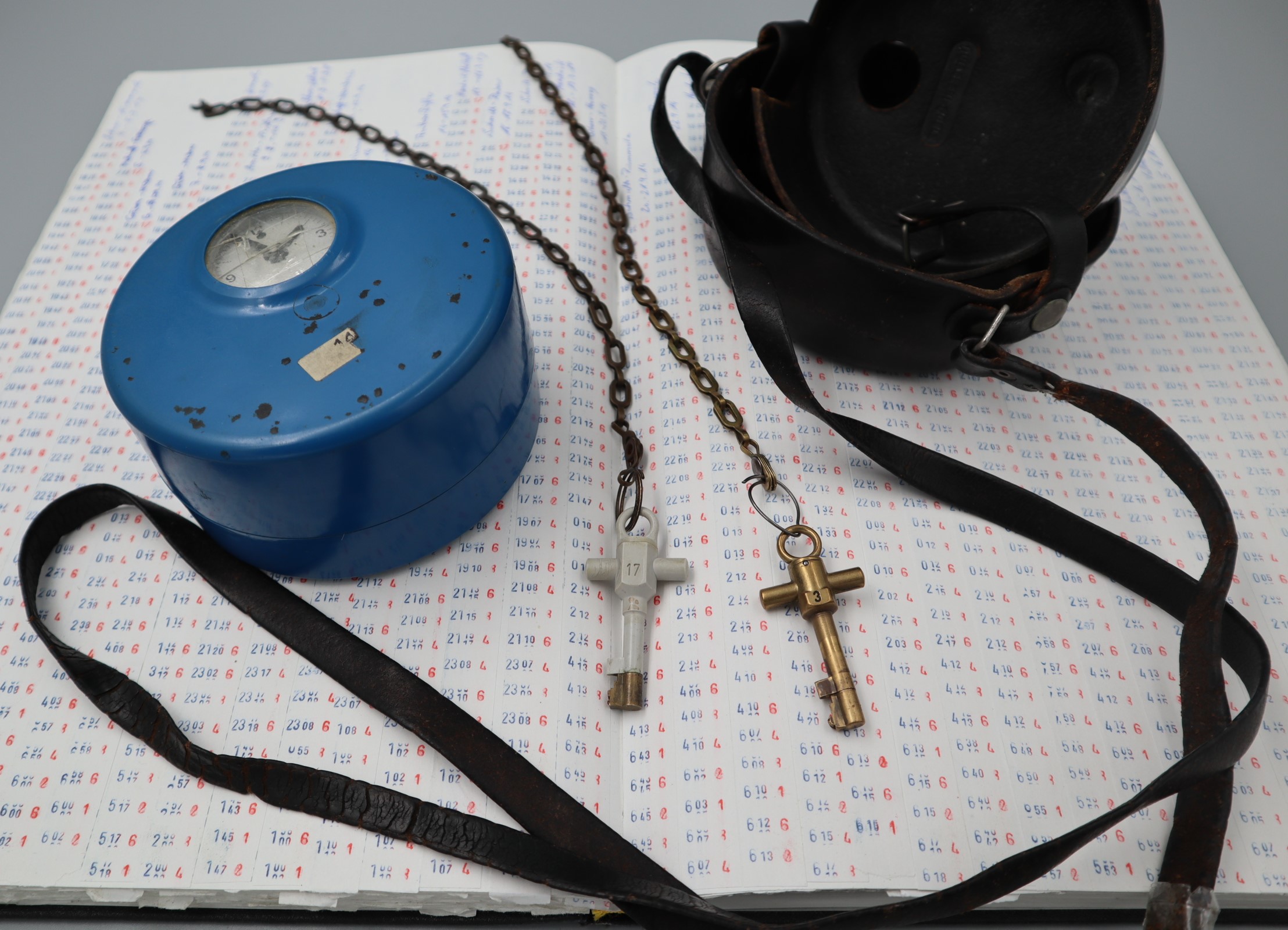 Blaues, hohes Uhrengehäuse, sogenannte Wächterkontrolluhr, die neben einem passenden Tragekoffer aus Leder und zwei Stockwerkschlüsseln auf einem Kontrollbuch liegt