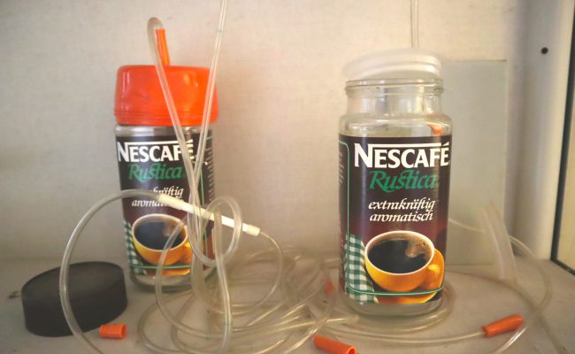 Destilationsapparatur aus zwei durchsichtigen Nescafé-Gläsern mit Löchern im Deckel, die über einen Schlauch miteinander verbunden sind, als Objekt im Strafvollzugsmuseum ausgestellt