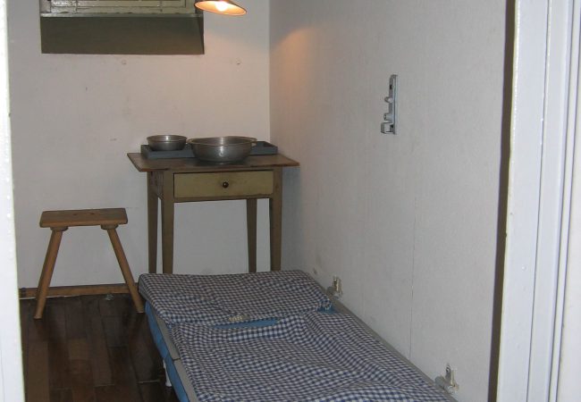 Blick in eine geöffnete Haftzelle mit Holzdielenboden, weißen Wänden, Wasserklosett, hochschließbarem Bett mit blaukarierter Bettwäsche und einem kleinen Holztisch mit zwei Schüsseln und einem Holzstuhl vor dem Fenster